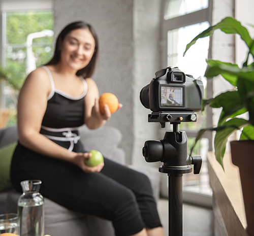 femme souriante en second-plan jouant avec des fruits et se filmant grâce à un appareil photo au premier-plan